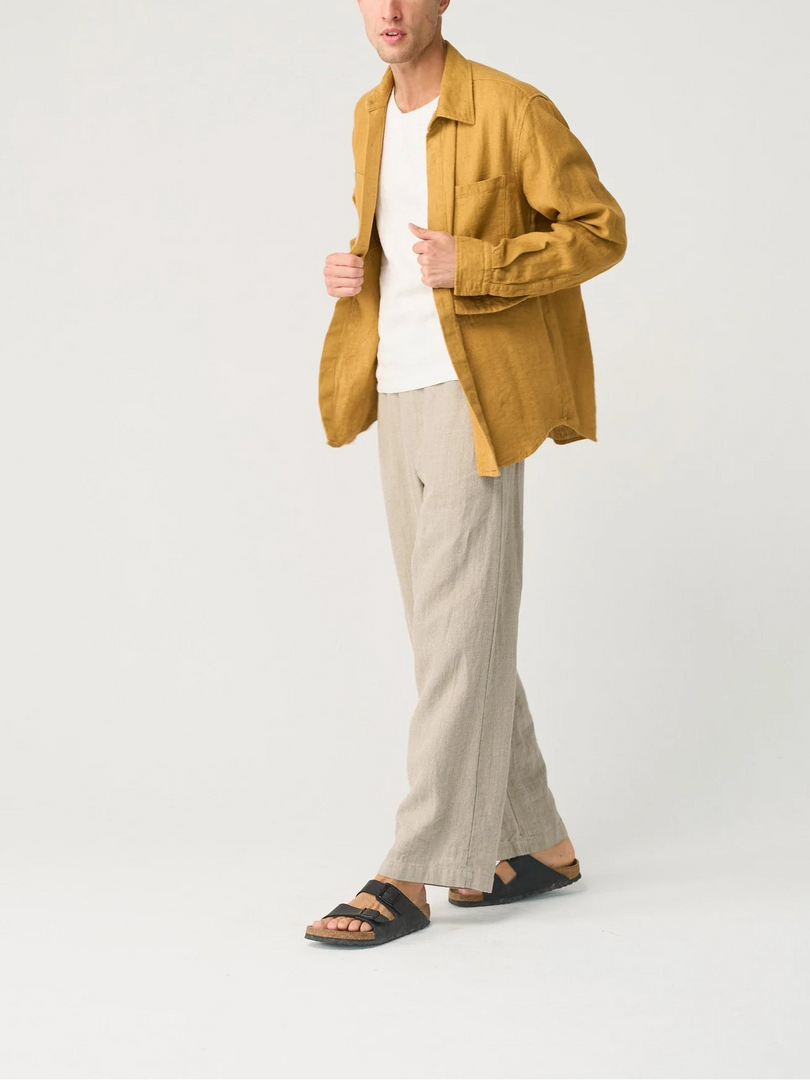 Man wearing a mustard jacket, white shirt, beige pants, and sandals on a white background